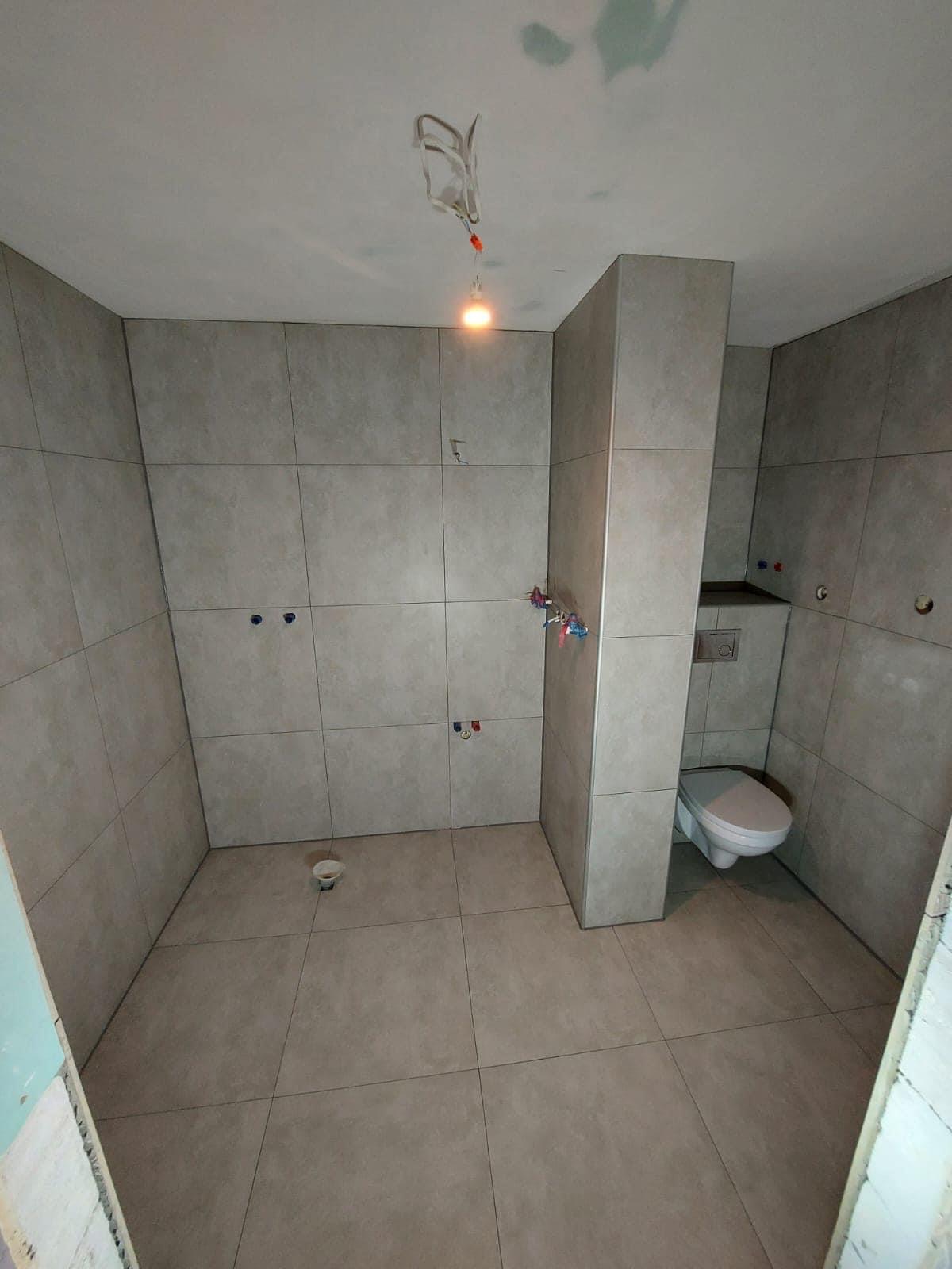 Łazienka w trakcie wykańczania: ułożone płytki ścienne i podłogowe w odcieniach szarości, widoczne instalacje wodne, podwieszana toaleta, surowe oświetlenie budowlane.