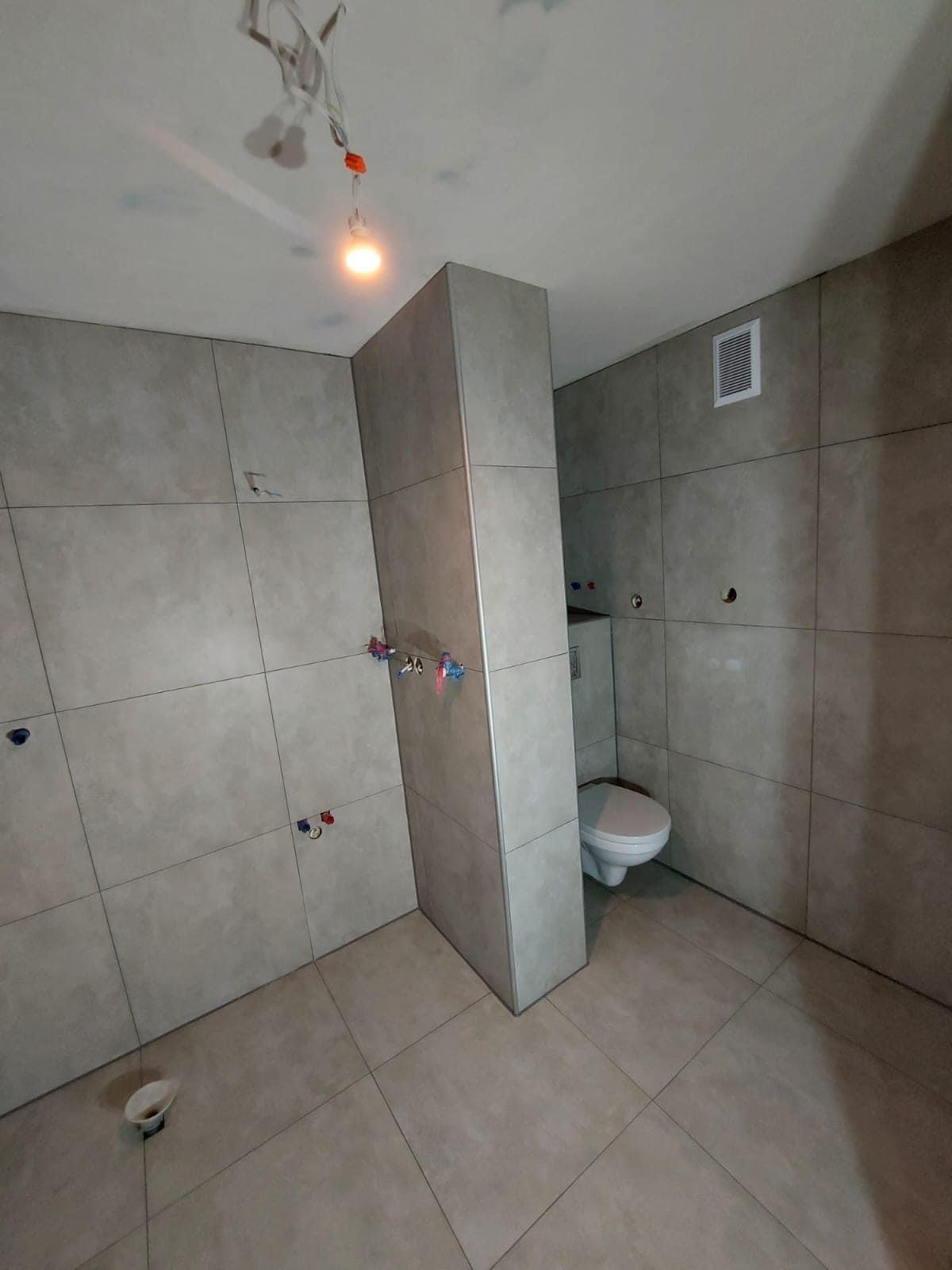 Wnętrze łazienki w trakcie remontu, widoczne szare płytki na ścianach i podłodze, częściowo zamontowana toaleta, wystające przewody elektryczne i niedokończone przyłącza wodne.