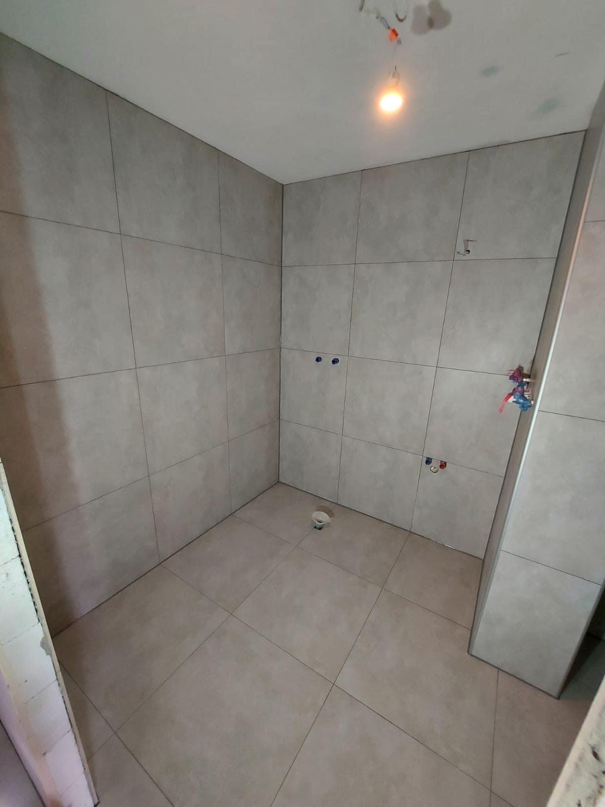 Wnętrze łazienki w trakcie remontu, widoczne płytki na ścianach i podłodze, instalacja wodna i elektryczna przygotowana, kubek na podłodze.