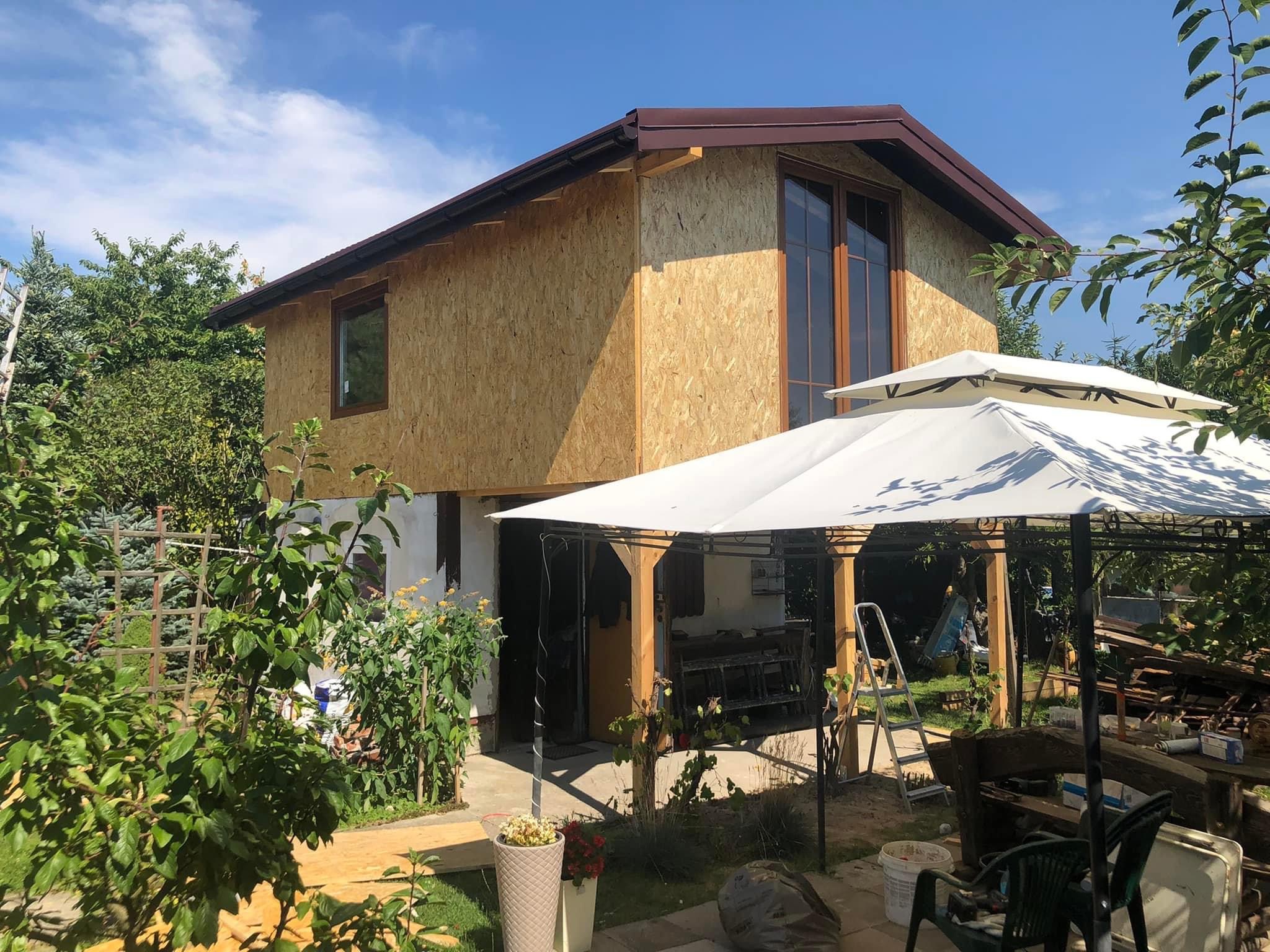 Dom w trakcie ocieplania, ściany pokryte płytami OSB, widoczne okna z brązowymi ramami, w tle błękitne niebo i zieleń drzew, w pierwszym planie altana ogrodowa z białym dachem.