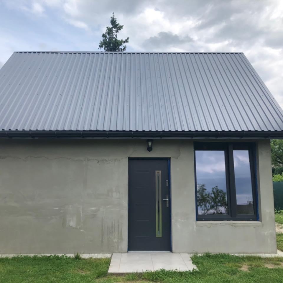 Niewykończony budynek z szarą blachodachówką, szarą elewacją przed położeniem tynku, ciemnymi drzwiami z pionowym przeszkleniem i oknem z widocznym odbiciem drzewa.