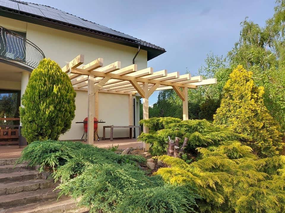 Nowoczesna drewniana pergola tarasowa z jasnego drewna, z widocznym grillem i małym stolikiem, otoczona bujną zielenią i krzewami, na tle jasnego domu z panelami słonecznymi na dachu.
