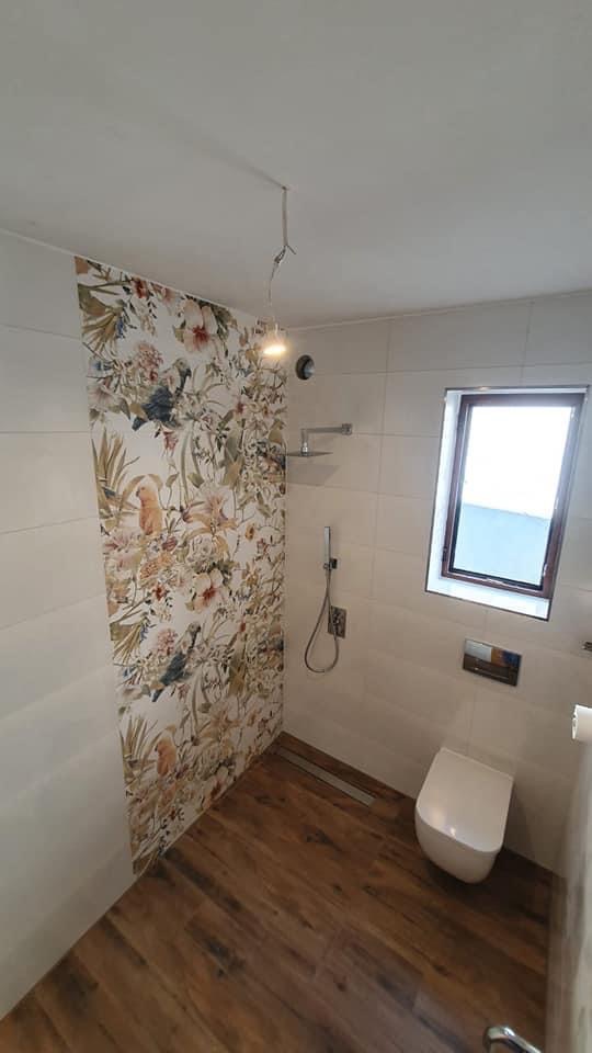 Wykończona łazienka z drewnopodobną podłogą, białą ceramiką i ścianą z motywem kwiatowym i papugami, widoczny prysznic z deszczownicą i oknem.