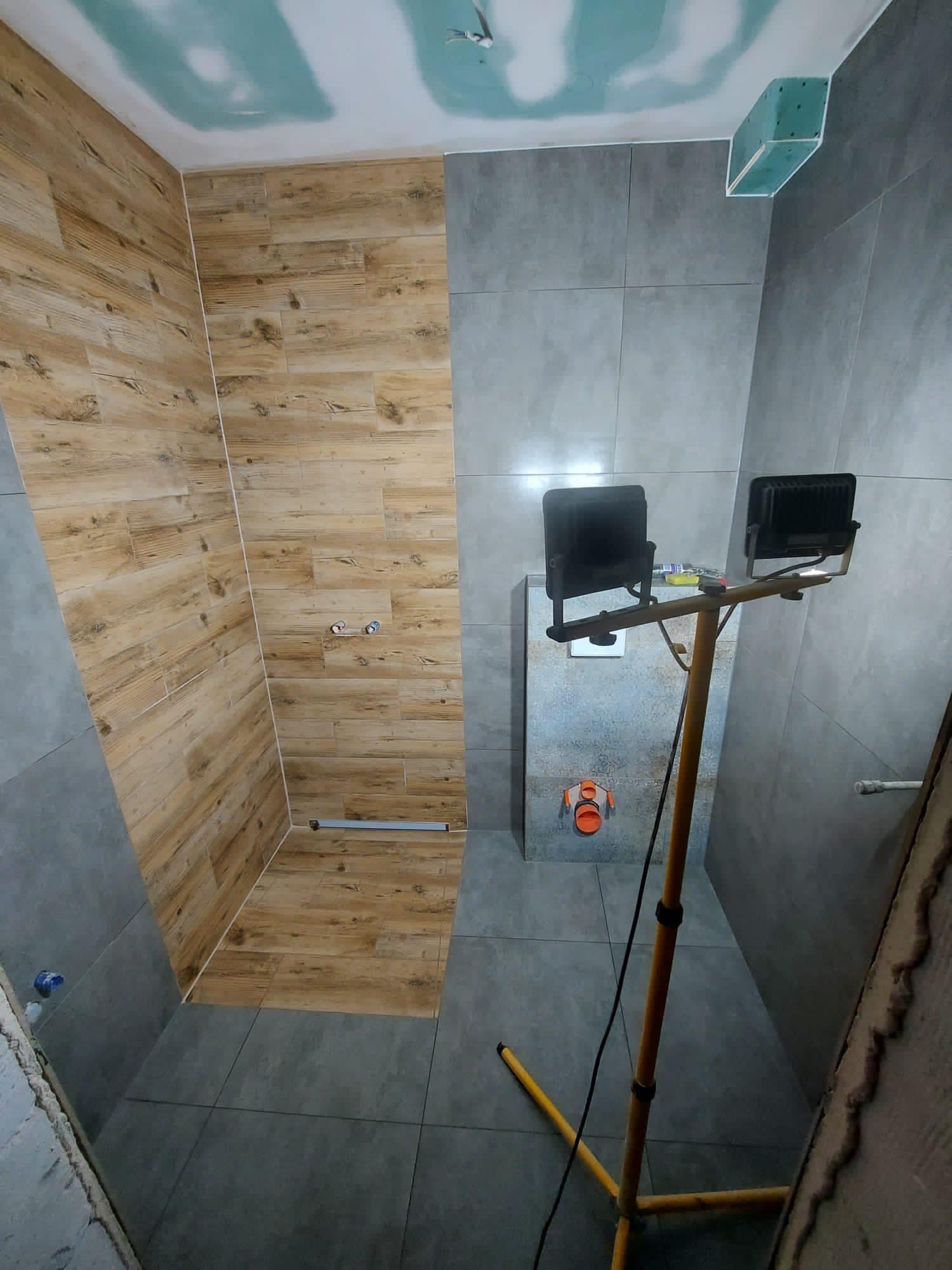 Wnętrze łazienki w trakcie remontu: ściany wyłożone szarymi i drewnopodobnymi płytkami, widoczna instalacja sanitarna i reflektor na statywie.