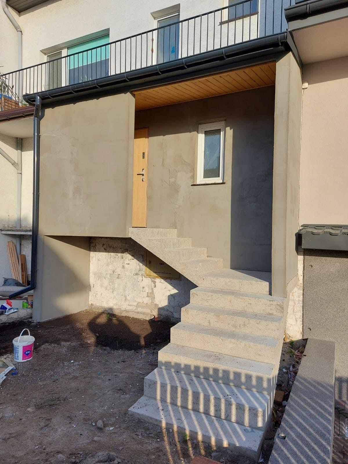 Nowe, betonowe schody prowadzące do wejścia budynku, ściany w trakcie szpachlowania, widoczny cień osoby fotografującej.