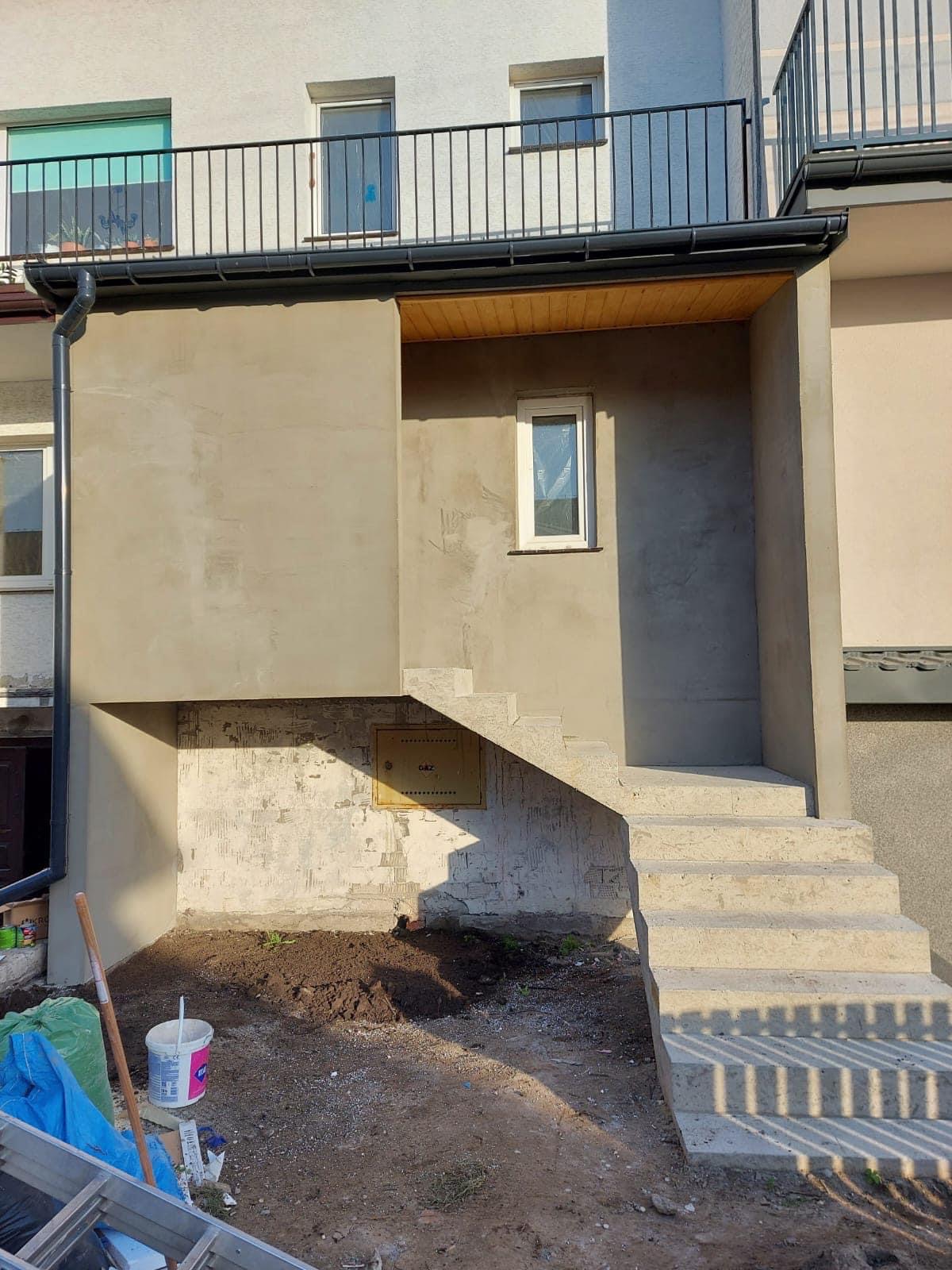 Narożnik budynku po szpachlowaniu, widoczne surowe, betonowe schody wejściowe, fragment elewacji z oknem i skrzynką gazową, narzędzia i materiały budowlane w tle.