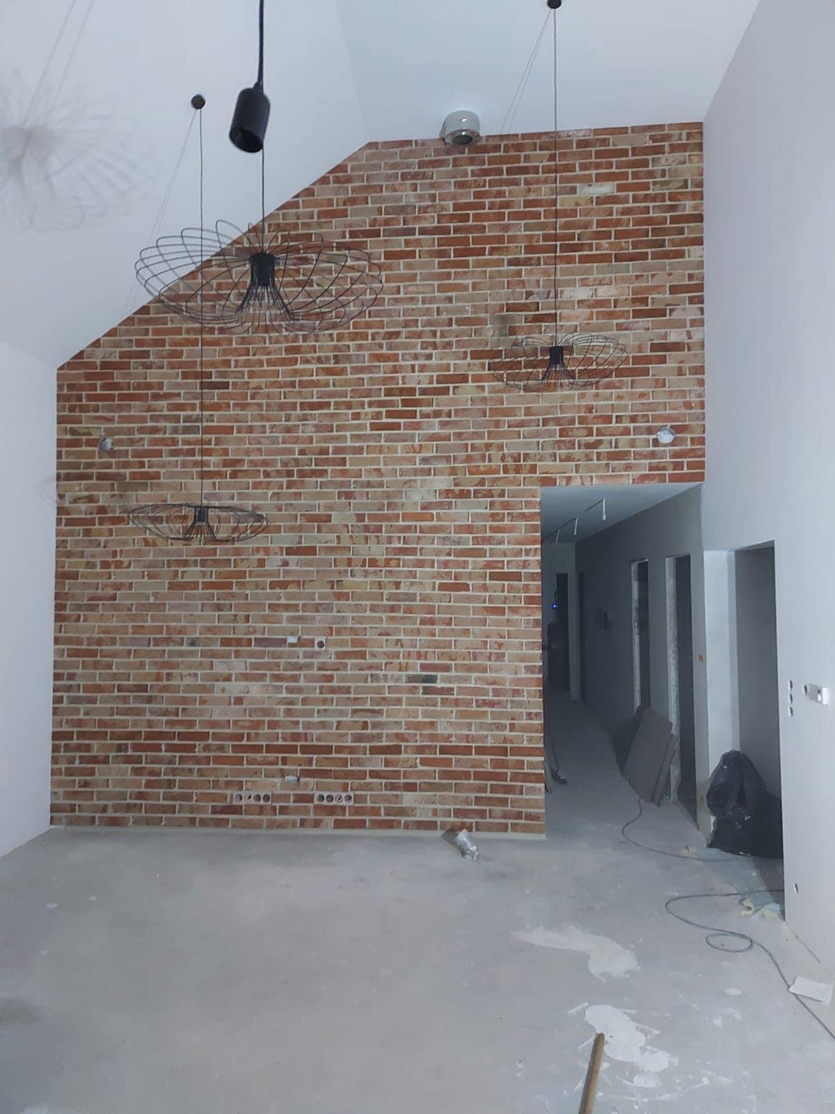 Surowa ceglana ściana z trzema nowoczesnymi lampami wiszącymi w przestronnym, jasnym wnętrzu z surową podłogą i otwartym przejściem do korytarza.