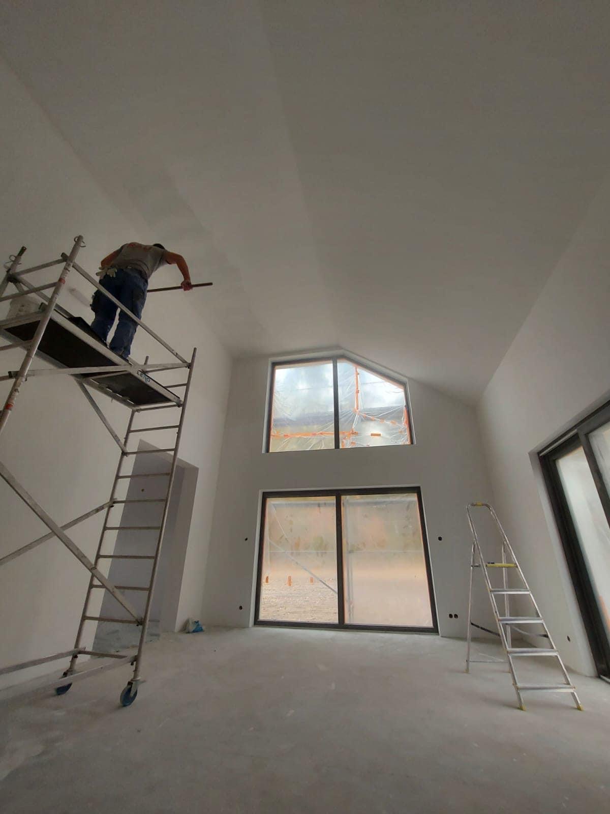 Malowanie sufitu w wysokim pomieszczeniu z dużymi oknami zabezpieczonymi folią, pracownik na rusztowaniu używa wałka, widoczna drabina. Sufit częściowo pomalowany.