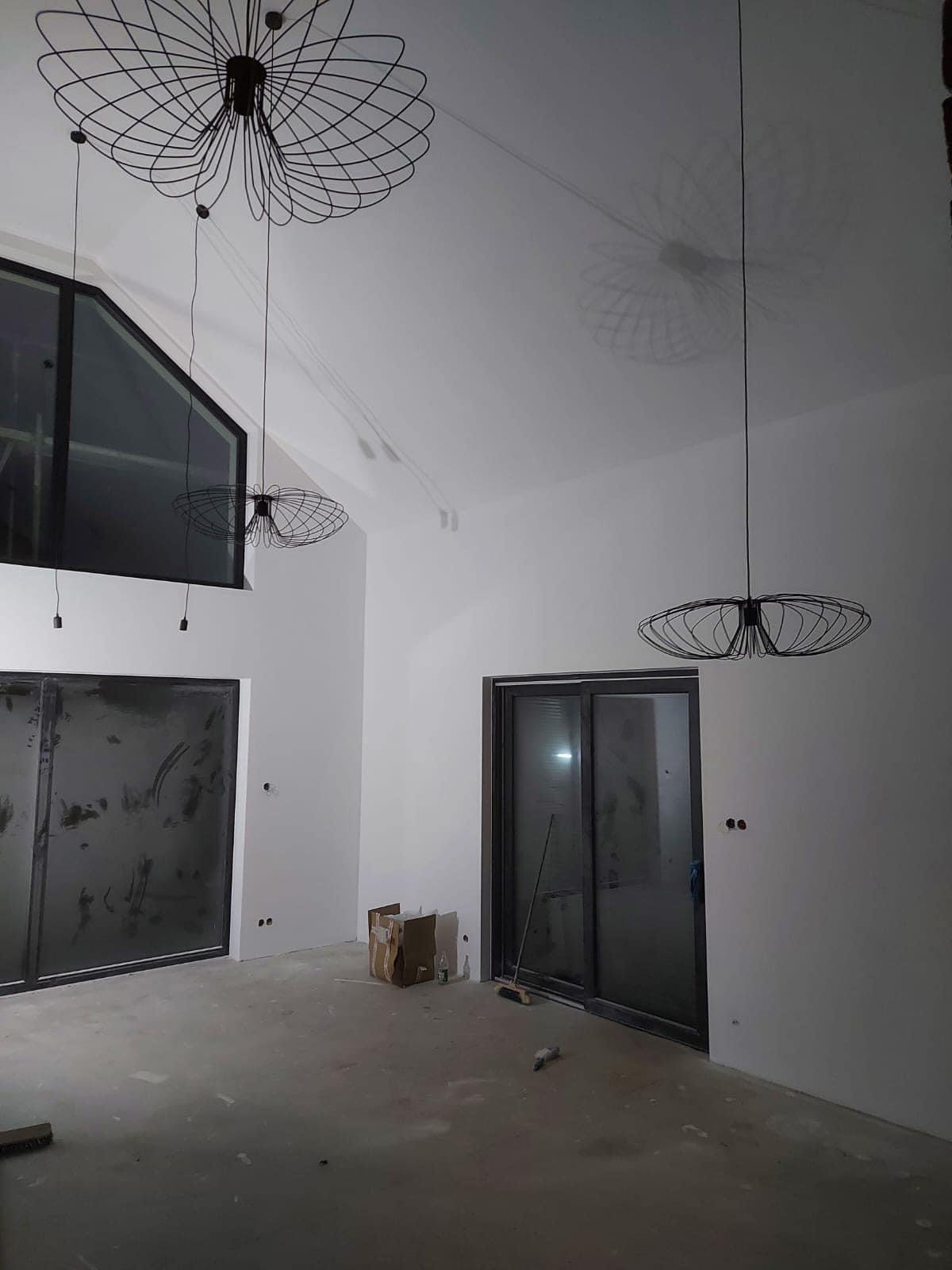 Wnętrze w trakcie wykańczania z trzema nowoczesnymi lampami wiszącymi o ażurowych kloszach, rzucającymi cienie na gładką, białą ścianę, widoczne surowe podłoże i okna z ciemnymi ramami.