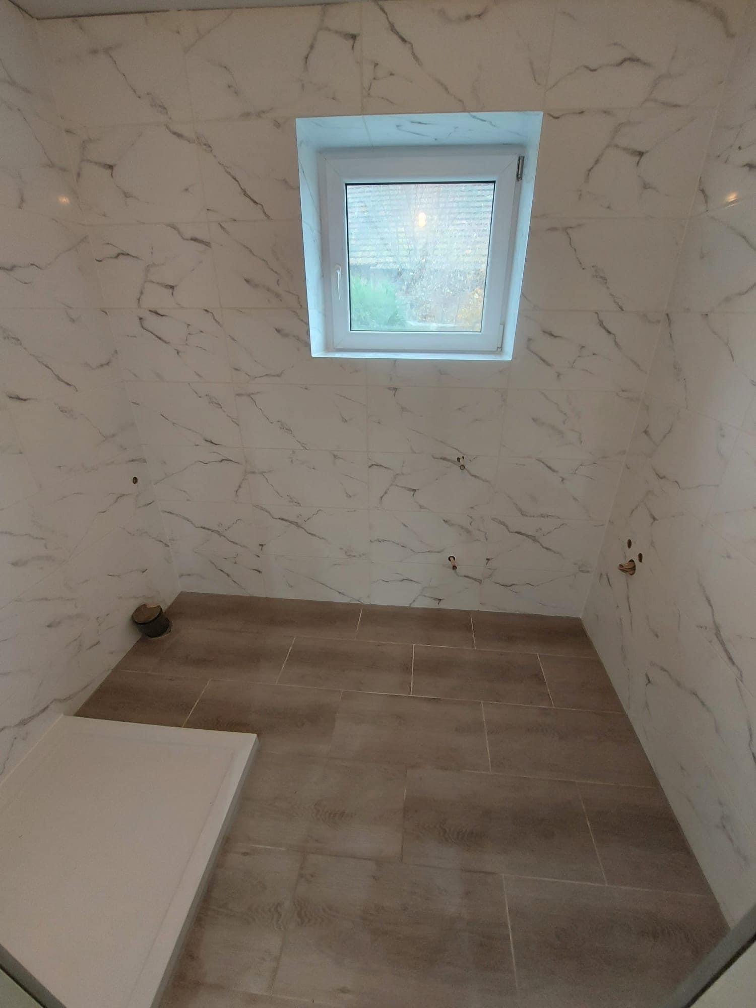 Wykończona łazienka na poddaszu z oknem dachowym, ściany wyłożone płytkami imitującymi marmur, podłoga z płytek drewnopodobnych, widoczne przyłącza hydrauliczne na ścianach i brodzik w rogu.