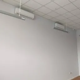 Wnętrze biurowe z zamontowanymi klimatyzatorami ściennymi i lampami sufitowymi, widoczne okablowanie i pusta przestrzeń.