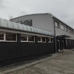 Nowoczesny budynek komercyjny z szarą elewacją i czarną cegłą, widoczny system wentylacyjny na dachu, pochmurne niebo w tle.