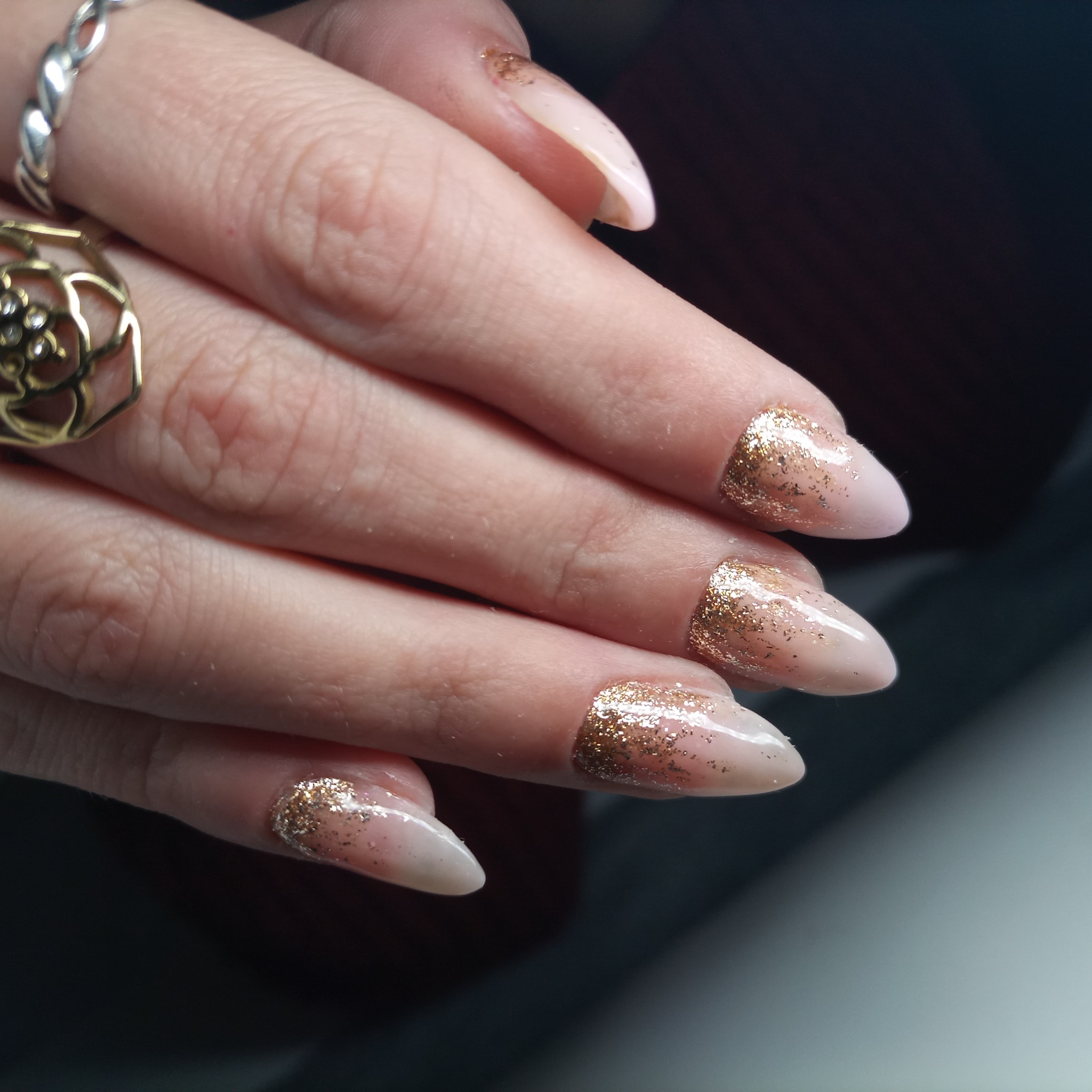 Dłoń z migdałowymi paznokciami pokrytymi jasnoróżowym lakierem i złotym brokatem na końcach, ozdobiona pierścionkami.