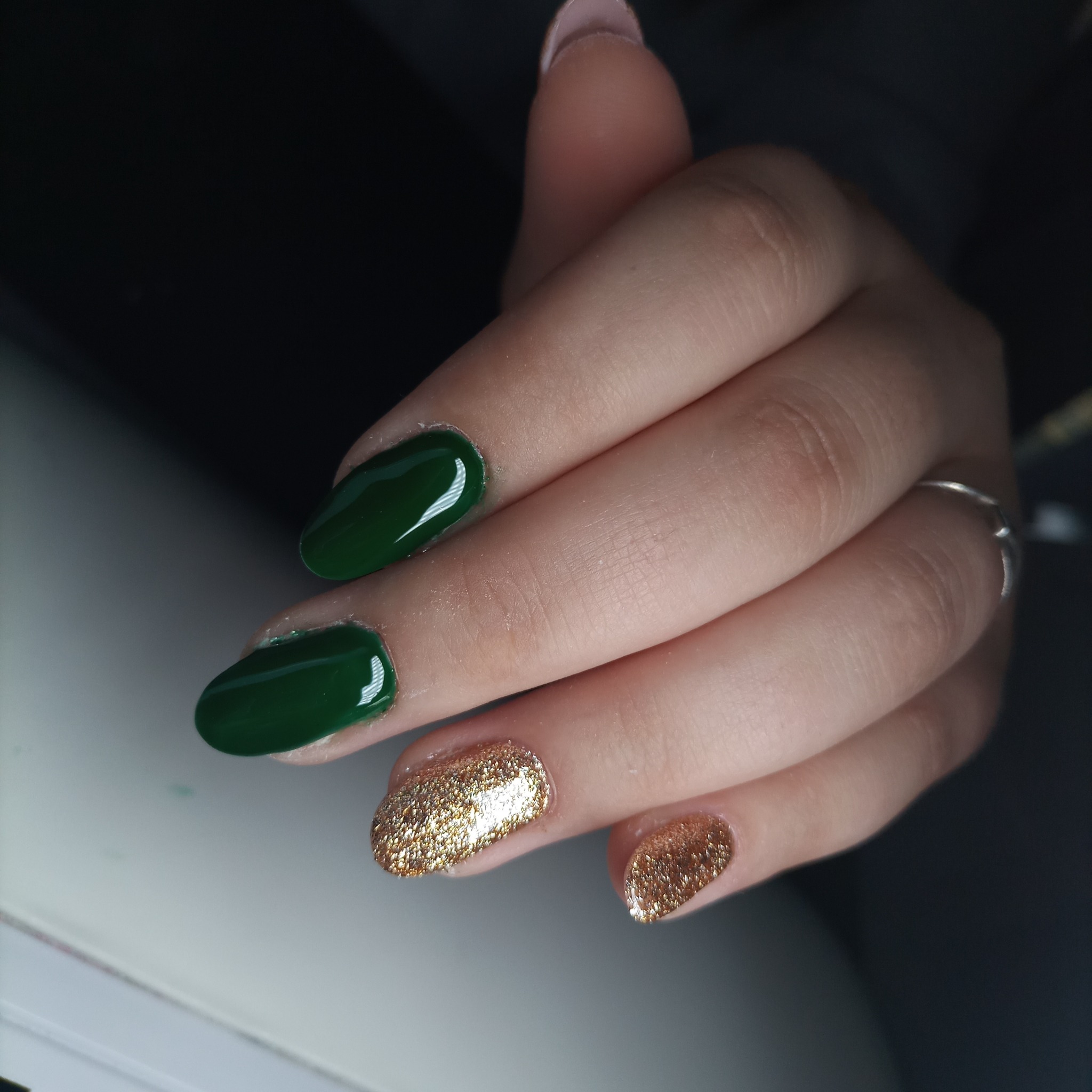 Dłoń z ciemnozielonymi paznokciami o owalnym kształcie i brokatowymi złotymi akcentami na dwóch palcach, widoczny srebrny pierścionek.
