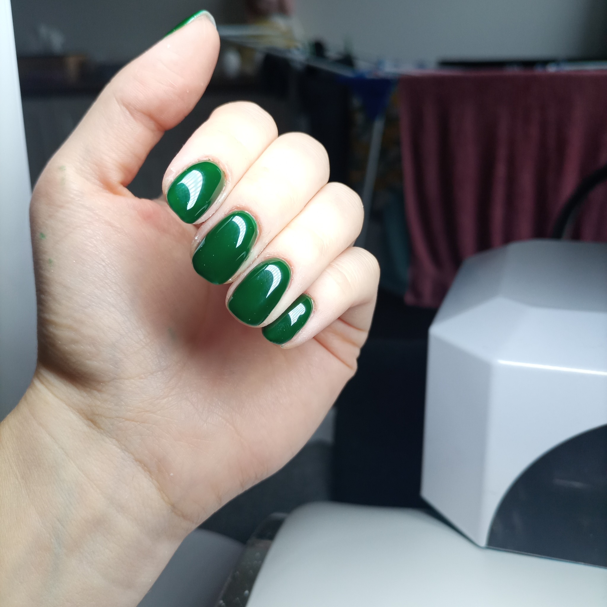 Dłoń z krótkimi, zaokrąglonymi paznokciami pomalowanymi na ciemnozielony, błyszczący lakier; w tle lampa UV do utwardzania lakieru hybrydowego.
