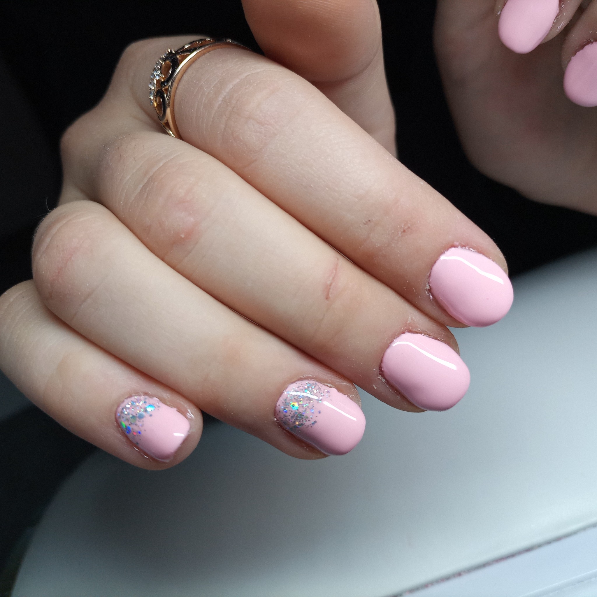 Ręka z delikatnym, różowym manicure hybrydowym; dwa paznokcie ozdobione srebrnym brokatem na końcach, złoty pierścionek z cyrkoniami na palcu wskazującym.