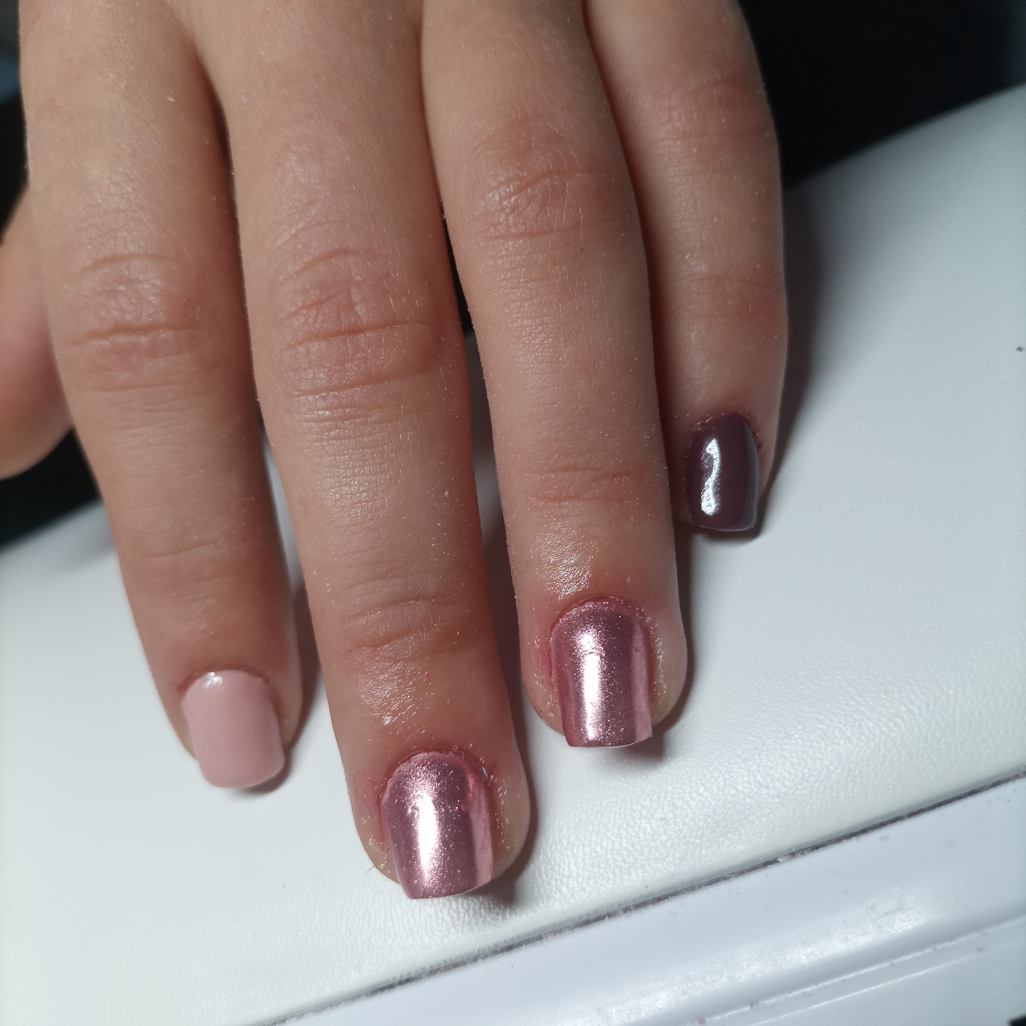 Dłoń z pomalowanymi paznokciami w odcieniach różu, metalicznego różu i brązu na białym tle.