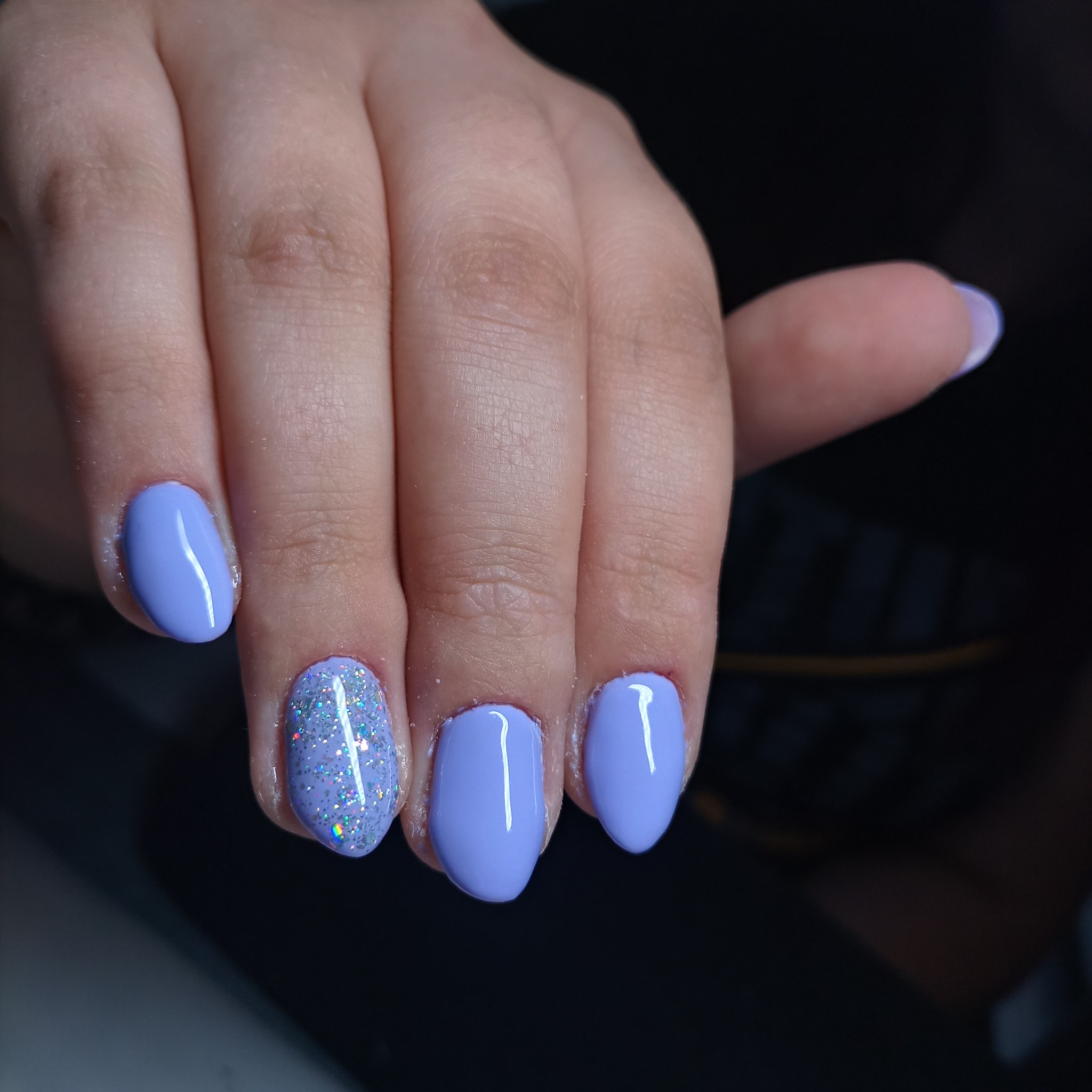 Dłoń z pomalowanymi na liliowo paznokciami, jeden z nich pokryty brokatem z efektem holo.