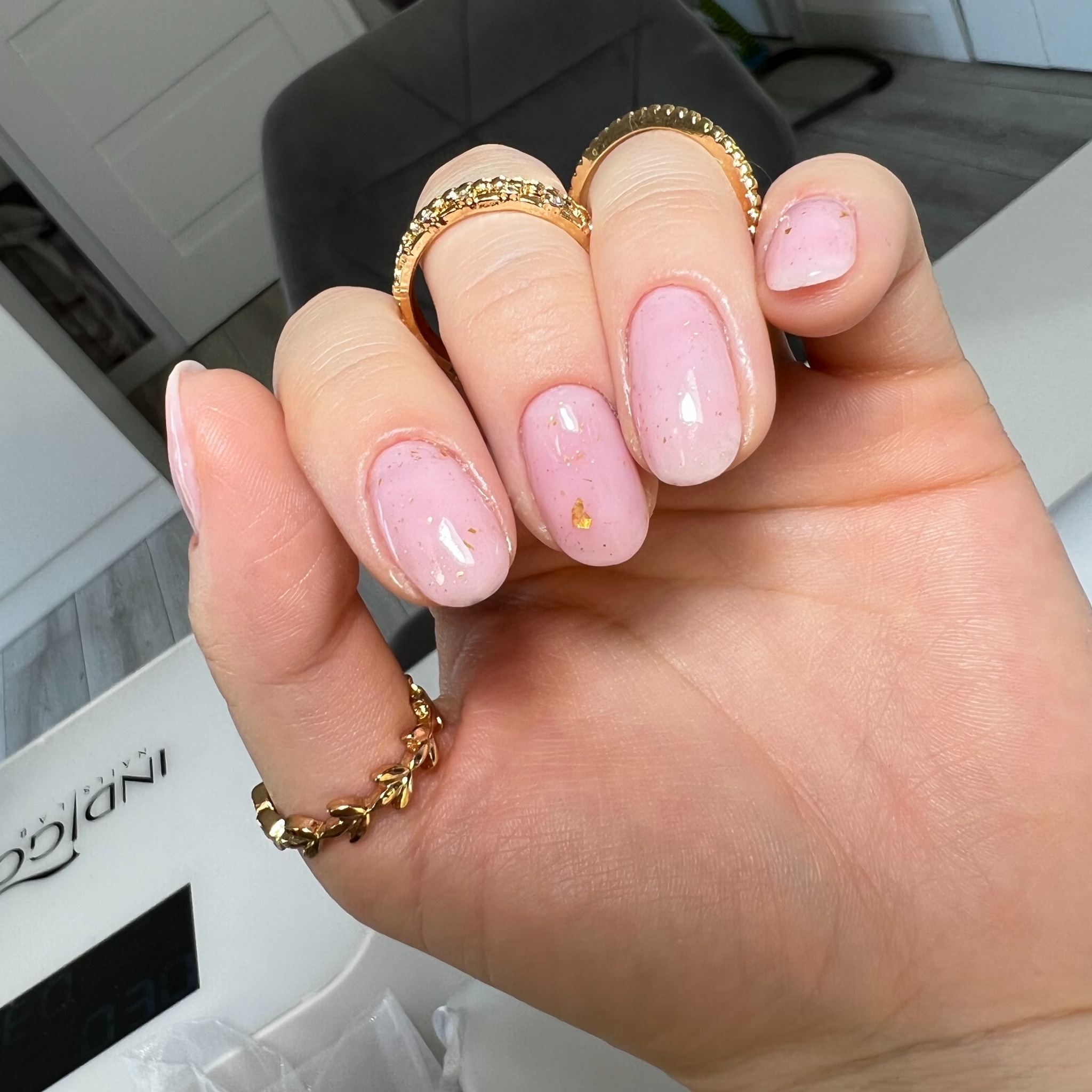 Dłoń z delikatnym, różowym manicure hybrydowym z drobnymi, złotymi płatkami, ozdobiona złotymi pierścionkami, na tle lampy UV do paznokci.