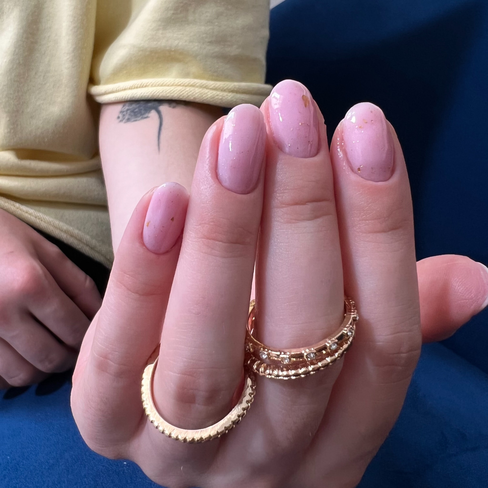 Delikatny, różowy manicure z drobnymi złotymi płatkami, ozdobiony dwoma złotymi pierścionkami na palcu wskazującym i środkowym, na tle granatowego materiału.