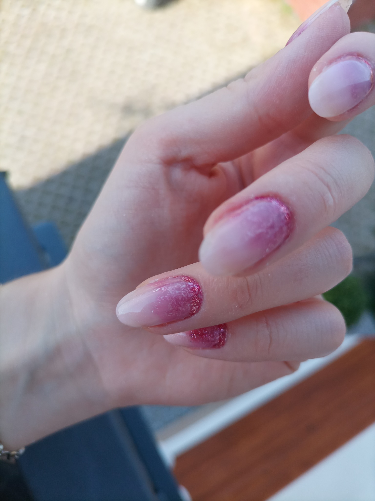 Dłoń z owalnymi paznokciami w odcieniach różu i bieli, z brokatowym wykończeniem przy skórkach, sfotografowana w dziennym świetle na zewnątrz.