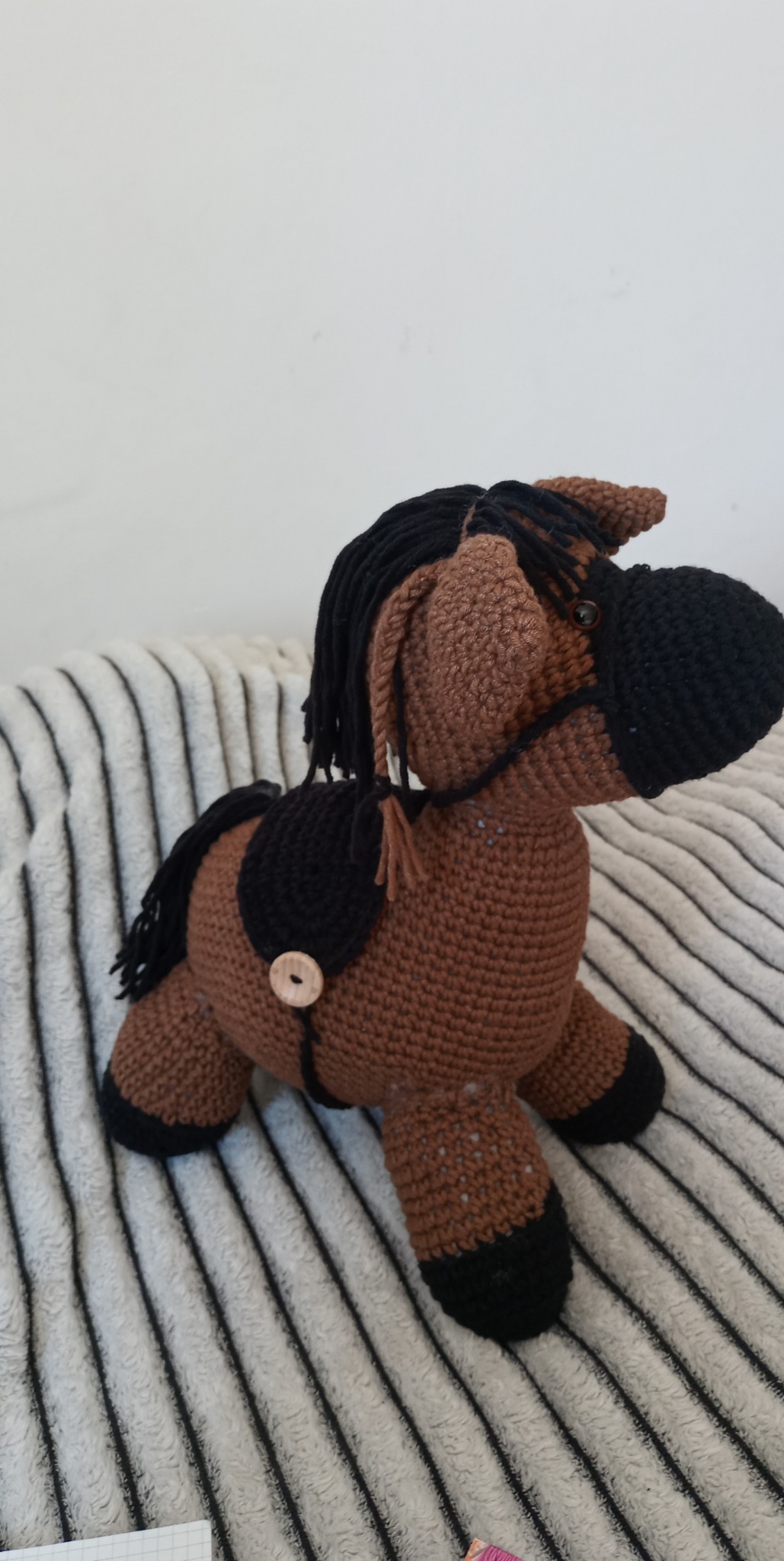 Ręcznie robiony, brązowo-czarny koń na biegunach z włóczki, z ciemną grzywą i ogonem, ozdobiony drewnianym guzikiem, leżący na puszystym, pasiastym kocu.