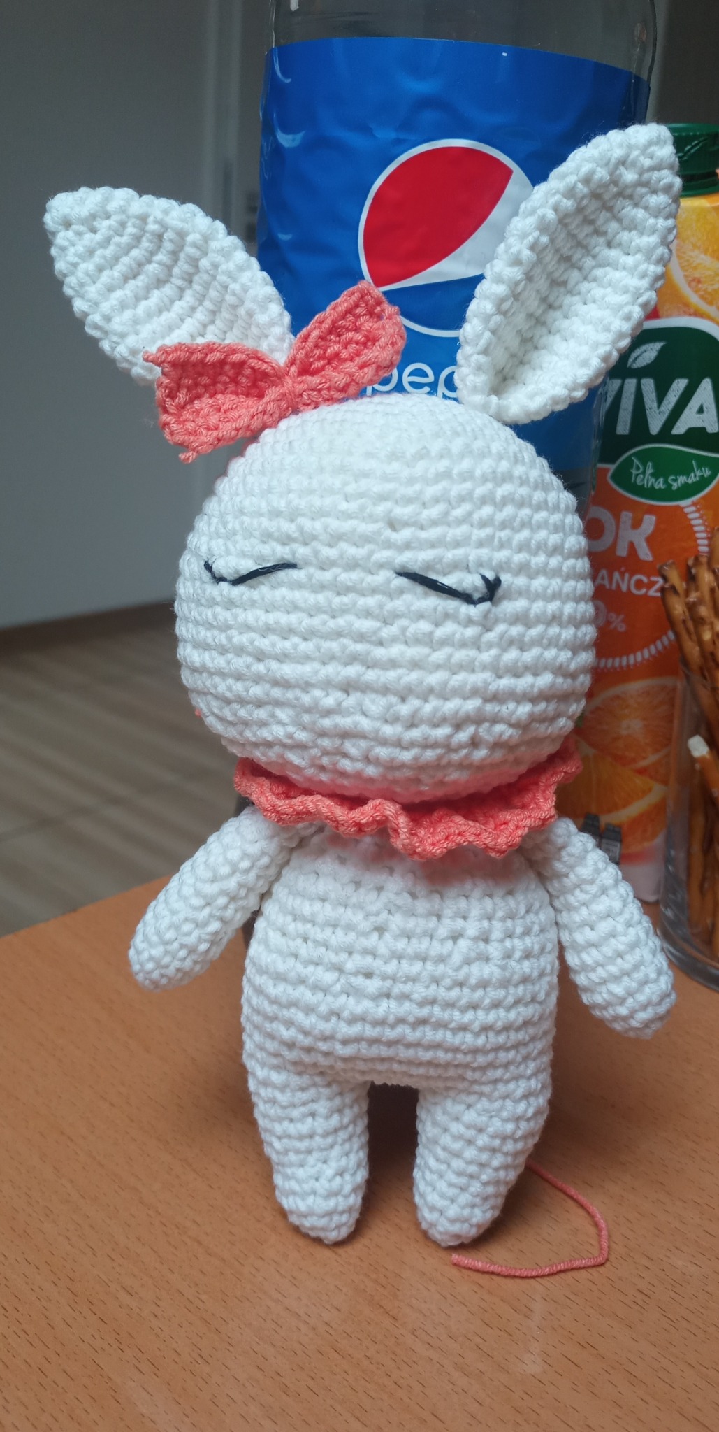 Ręcznie robiona, biała, szydełkowa maskotka królika z koralową kokardką i falbanką na szyi, stojąca na drewnianym stole, z napojami w tle.