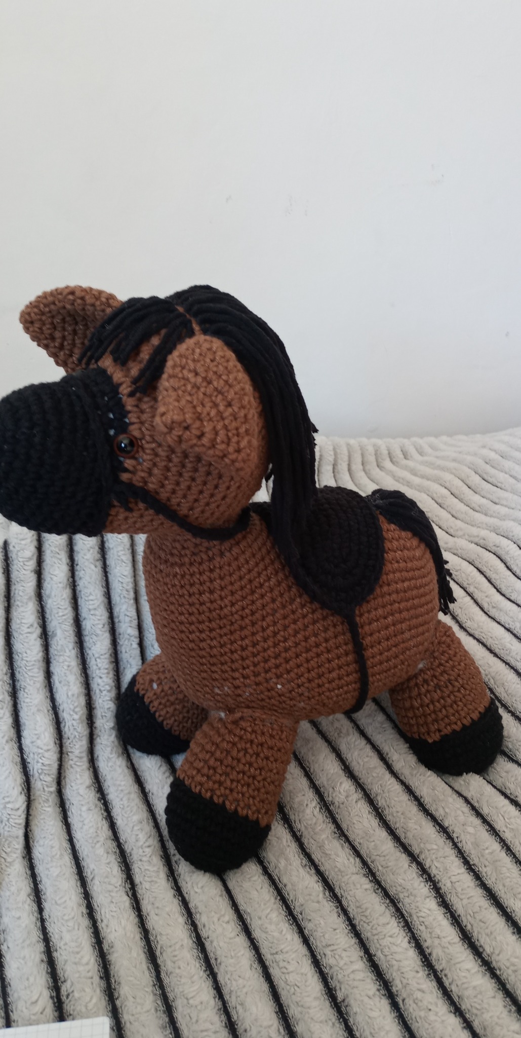 Ręcznie robiony, brązowy, pluszowy koń z czarną grzywą i kopytami leży na puszystej, biało-szarej, prążkowanej tkaninie.