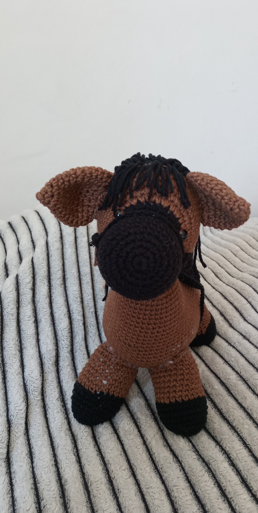 Brązowy, ręcznie robiony na szydełku koń z czarną grzywą i kopytami, siedzący na biało-czarnym pasiastym tle.