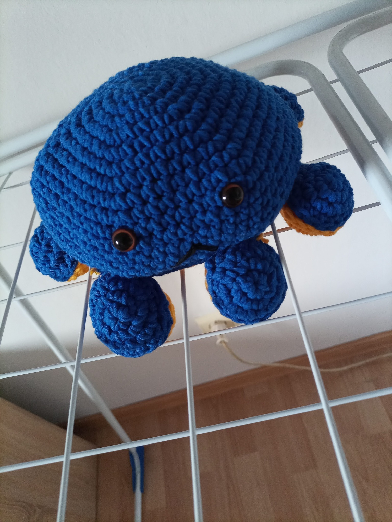 Niebieski, ręcznie robiony, pluszowy ośmiornica z żółtymi akcentami, zawieszony na białej suszarce do ubrań w pomieszczeniu z drewnianą podłogą.