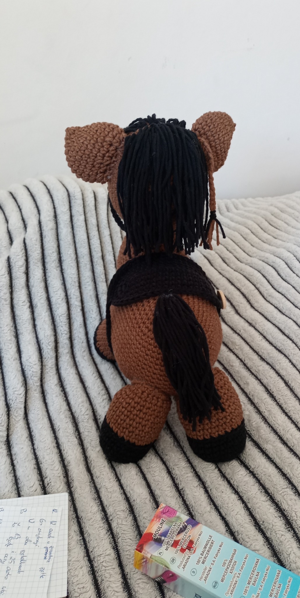 Tył brązowego, ręcznie robionego konika na biegunach z czarną grzywą i ogonem, leżącego na szarym, prążkowanym kocu. W tle widoczne notatki i pudełko nici.