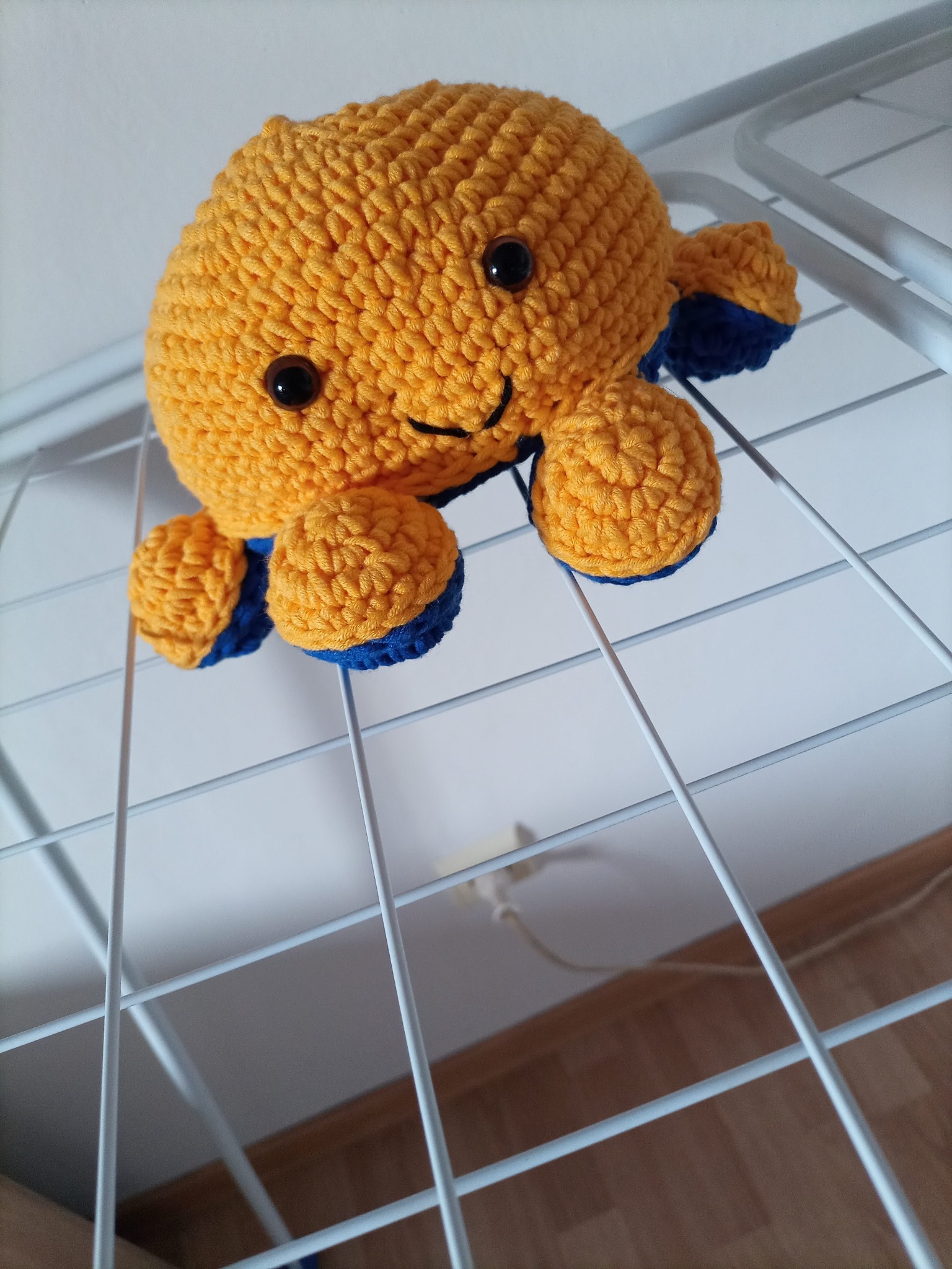 Żółto-niebieska, szydełkowa zabawka w kształcie ośmiornicy z czarnymi oczkami, umieszczona na metalowej suszarce do ubrań.