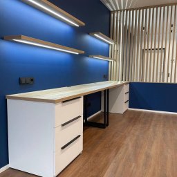 Berg Meble - Długi, drewniany blat biurka z białymi szafkami po bokach na tle ciemnoniebieskiej ściany z podświetlanymi półkami i drewnianej ażurowej ścianki działowej.