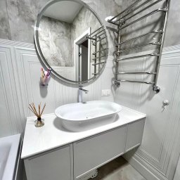 Berg Meble - Nowoczesna łazienka z owalną umywalką nablatową na białej szafce, okrągłym lustrem z podświetleniem i chromowanym grzejnikiem drabinkowym na tle marmurowych ścian.