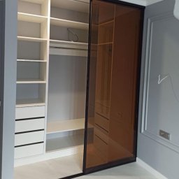 Berg Meble - Szafa wnękowa z przesuwnymi drzwiami z brązowego szkła, widoczne półki, szuflady i drążek na ubrania, nowoczesny design w szarym pomieszczeniu.