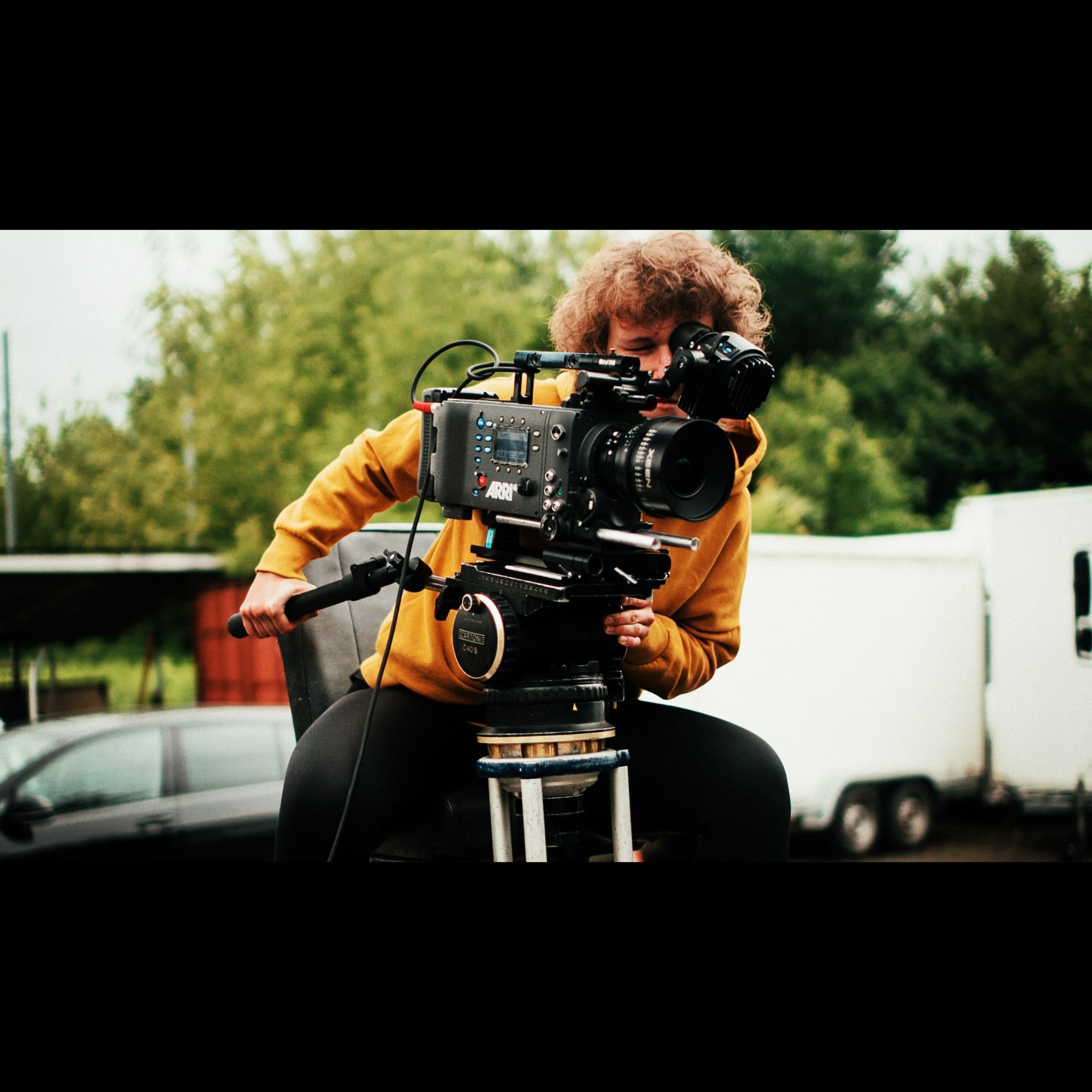 Osoba z kręconymi włosami obsługuje profesjonalną kamerę ARRI zamontowaną na statywie, ubrana w żółtą bluzę, w tle zieleń, samochód i przyczepa
