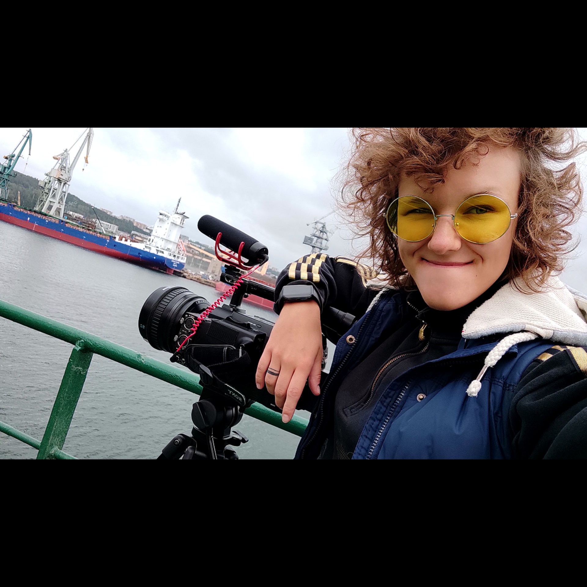 Kobieta z kręconymi włosami w żółtych okularach, uśmiechnięta, pozuje z kamerą i mikrofonem na tle portu z dźwigami i statkami.