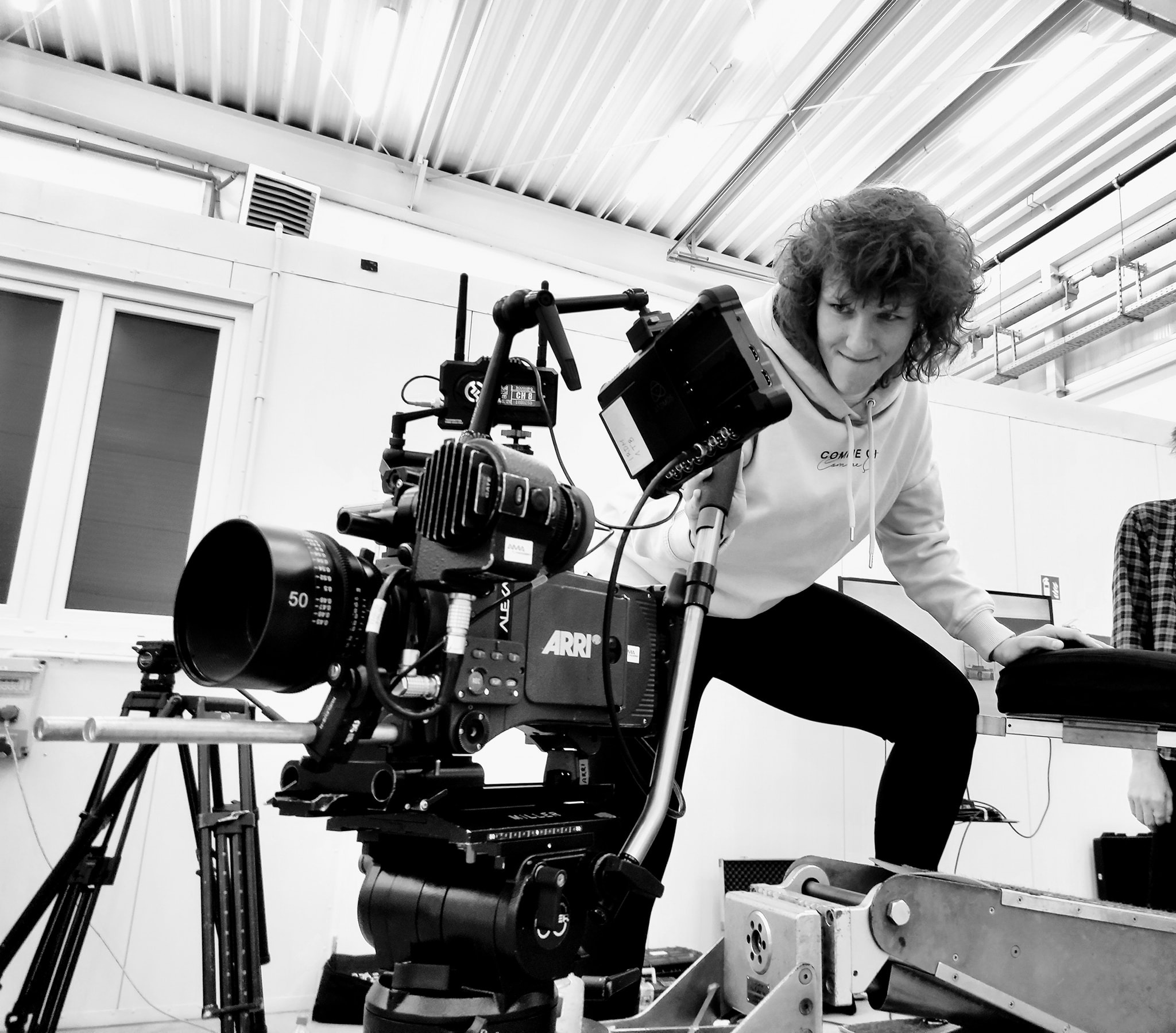 Czarno-białe zdjęcie przedstawia kobietę pochylającą się nad profesjonalną kamerą filmową ARRI Alexa z dużym obiektywem, umieszczoną na specjalistycznym wózku, w pomieszczeniu z oknami...