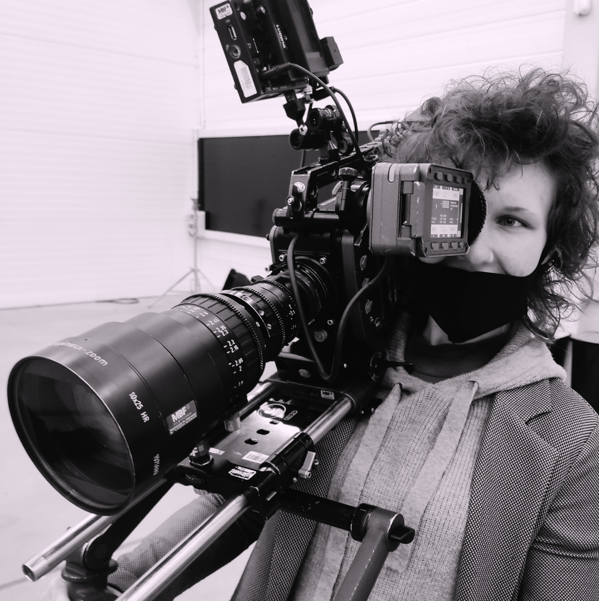 Kobieta z krótkimi kręconymi włosami, w masce, obsługująca profesjonalną kamerę filmową z obiektywem zoom 10x25 HR marki MBF Filtechnik, zamontowaną na statywie z szynami, w studio filmowym...