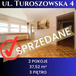 Joanna Ewa Balińska - Wnętrze 2-pokojowego mieszkania w Łodzi, 37,52 m², 3 piętro, ul. Turoszowska 4. Na zdjęciu widoczny napis 'SPRZEDANE' z czerwoną fajką. Parkiet na podłodze.