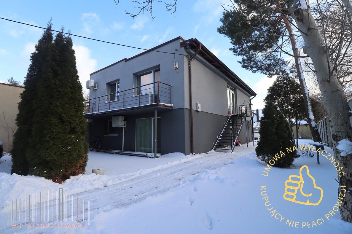 Dwukondygnacyjny budynek z balkonem i zewnętrznymi schodami w zimowej scenerii. Śnieg pokrywa trawnik i ścieżkę prowadzącą do wejścia. Widoczne drzewa i krzewy.