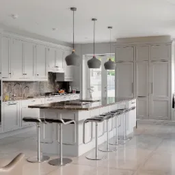 Jasna kuchnia w stylu klasycznym z wyspą z blatem z ciemnego marmuru, oświetlona trzema szarymi lampami wiszącymi. W tle zabudowa kuchenna z białymi szafkami i sprzętem AGD.