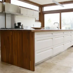 Nowoczesna kuchnia z wyspą: białe fronty szafek, drewniany blat i boczne panele wyspy, okno z drewnianą ramą, beżowa kamienna podłoga, widok na ogród.