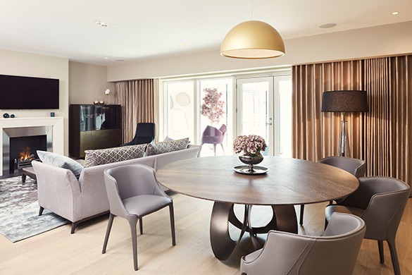 Jasne wnętrze apartamentu hotelowego z okrągłym stołem, szarymi krzesłami, sofą z poduszkami, kominkiem z płomieniami, telewizorem, złotą lampą wiszącą i zasłonami w odcieniach beżu.