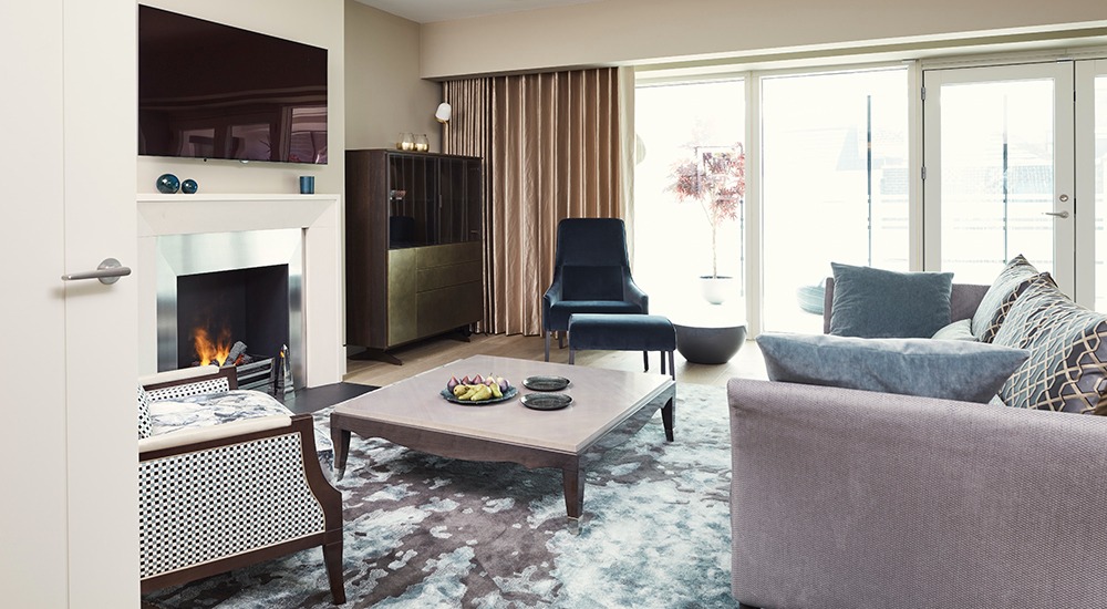 Salon w apartamencie hotelowym: kominek z paleniskiem, telewizor, szafka z dekoracjami, sofa z poduszkami, fotel z podnóżkiem, dywan z abstrakcyjnym wzorem, owoce na stoliku kawowym.