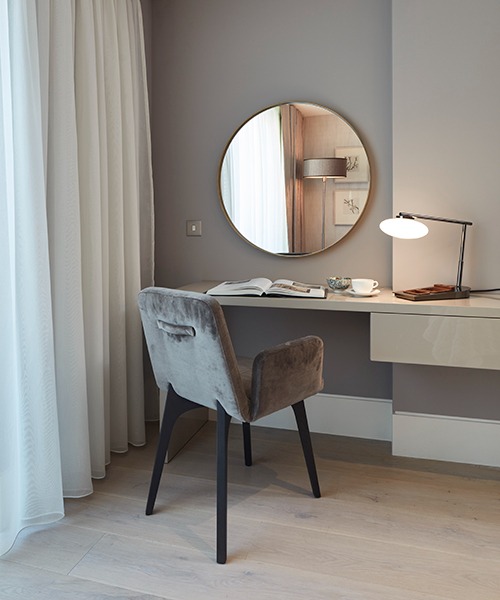 Apartament hotelowy: biurko z okrągłym lustrem, szarą tapicerowaną krzesłem, lampką, książką i filiżanką kawy, oświetlone naturalnym światłem z okna z białymi zasłonami.