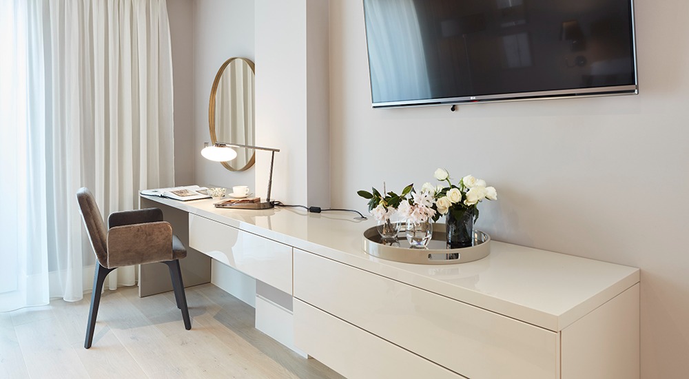 Apartament hotelowy w Dublinie: biurko z połyskiem, kwiaty w wazonie na srebrnej tacy, okrągłe lustro nad biurkiem, telewizor na ścianie, krzesło z brązowego weluru.