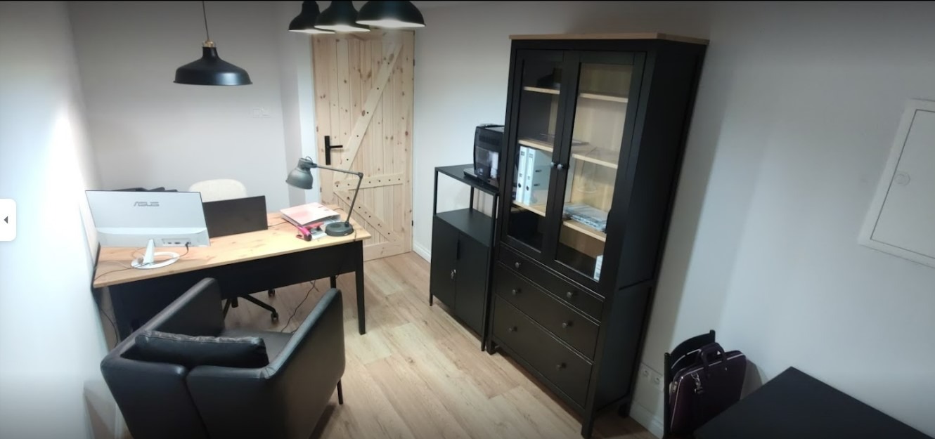 Jasne biuro z drewnianym biurkiem, czarnym krzesłem, regałem z szufladami i drewnianymi drzwiami w stylu rustykalnym.