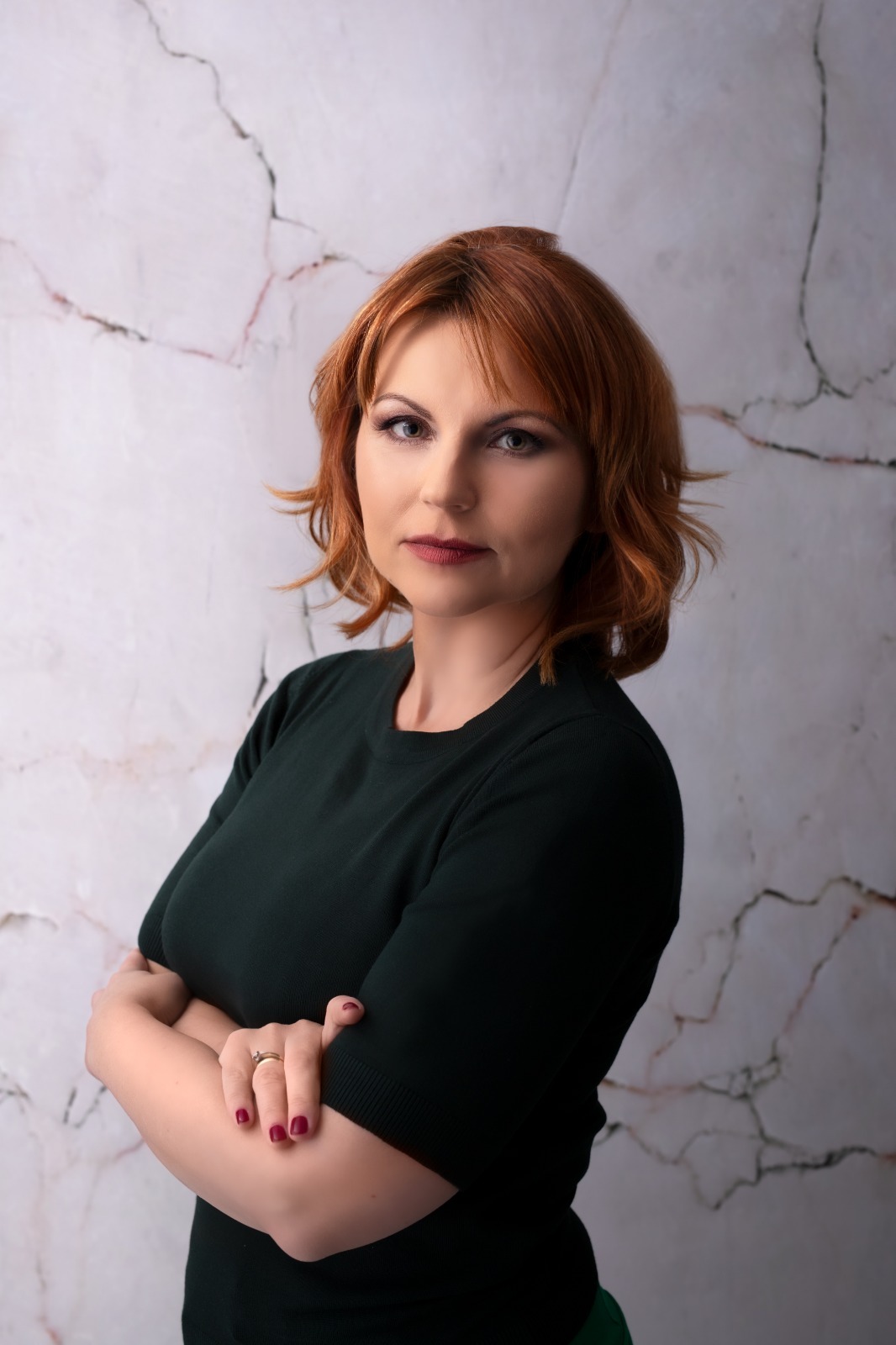 Portret kobiety z rudymi włosami w ciemnozielonej bluzce na tle marmurowej ściany w Lublinie, z założonymi rękami i pierścionkiem na palcu.