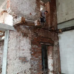 Pracownik z wiertarką udarową w otworze w ceglanej ścianie podczas prac remontowych we wnętrzu budynku.